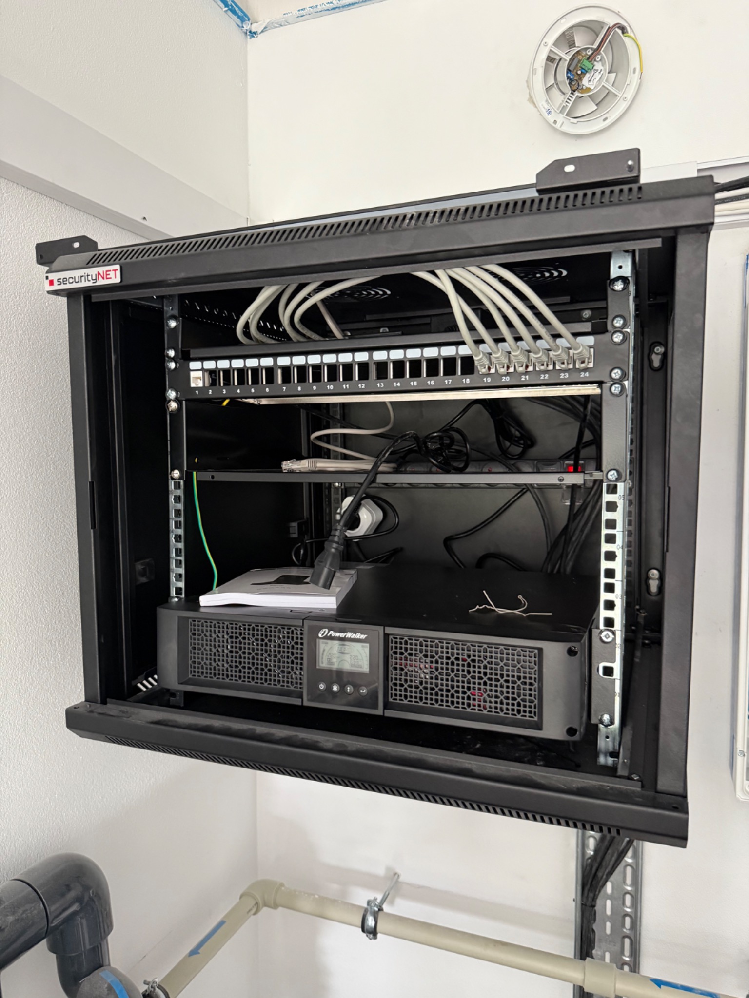 Szafa rack securityNET z patch panelem, switchem i zasilaczem awaryjnym PowerWalker. Widoczne kable patchcord i przewody. Na ścianie biały czujnik.