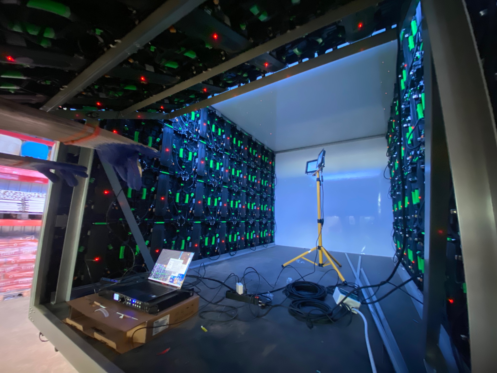 Instalacja modułowej ściany LED z widocznym laptopem i okablowaniem wewnątrz metalowej konstrukcji. Widoczne elementy oświetlenia roboczego i fragment magazynu.