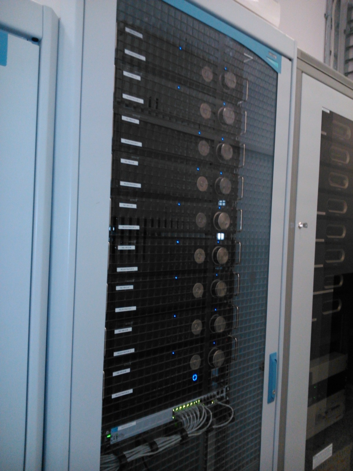Szafa serwerowa z panelami, okablowaniem i diodami LED, zamknięta w metalowej obudowie z siatkowymi drzwiami, widoczna w serwerowni.