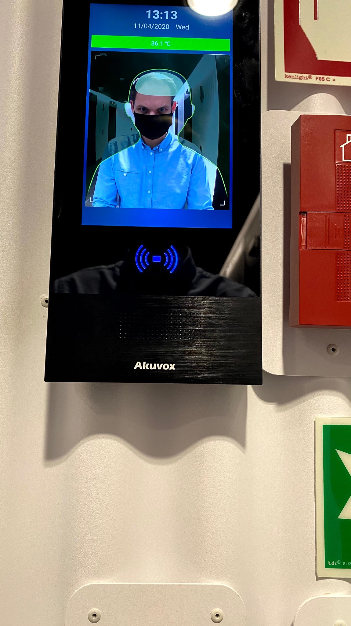 Terminal do pomiaru temperatury i rozpoznawania twarzy Akuvox, zamontowany na ścianie obok czerwonej skrzynki alarmowej. Na ekranie skan twarzy mężczyzny w maseczce.