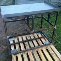 Stół roboczy ze stali nierdzewnej w trakcie budowy, umieszczony na drewnianej palecie, z widocznym ogrodzeniem betonowym w tle.