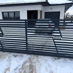 SaldaTech - Nowoczesna, pozioma brama wjazdowa z szarego metalu o geometrycznym wzorze na tle zasypanego śniegiem podjazdu i białego domu z garażem.