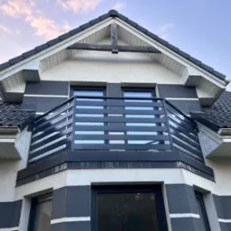 Nowoczesny balkon z poziomą, czarną balustradą na tle dwukolorowej elewacji domu z ciemną dachówką