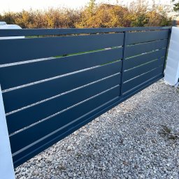 SaldaTech - Nowoczesna, przesuwna brama w kolorze grafitowym z poziomymi panelami, osadzona między białymi, murowanymi słupkami na żwirowym podłożu.