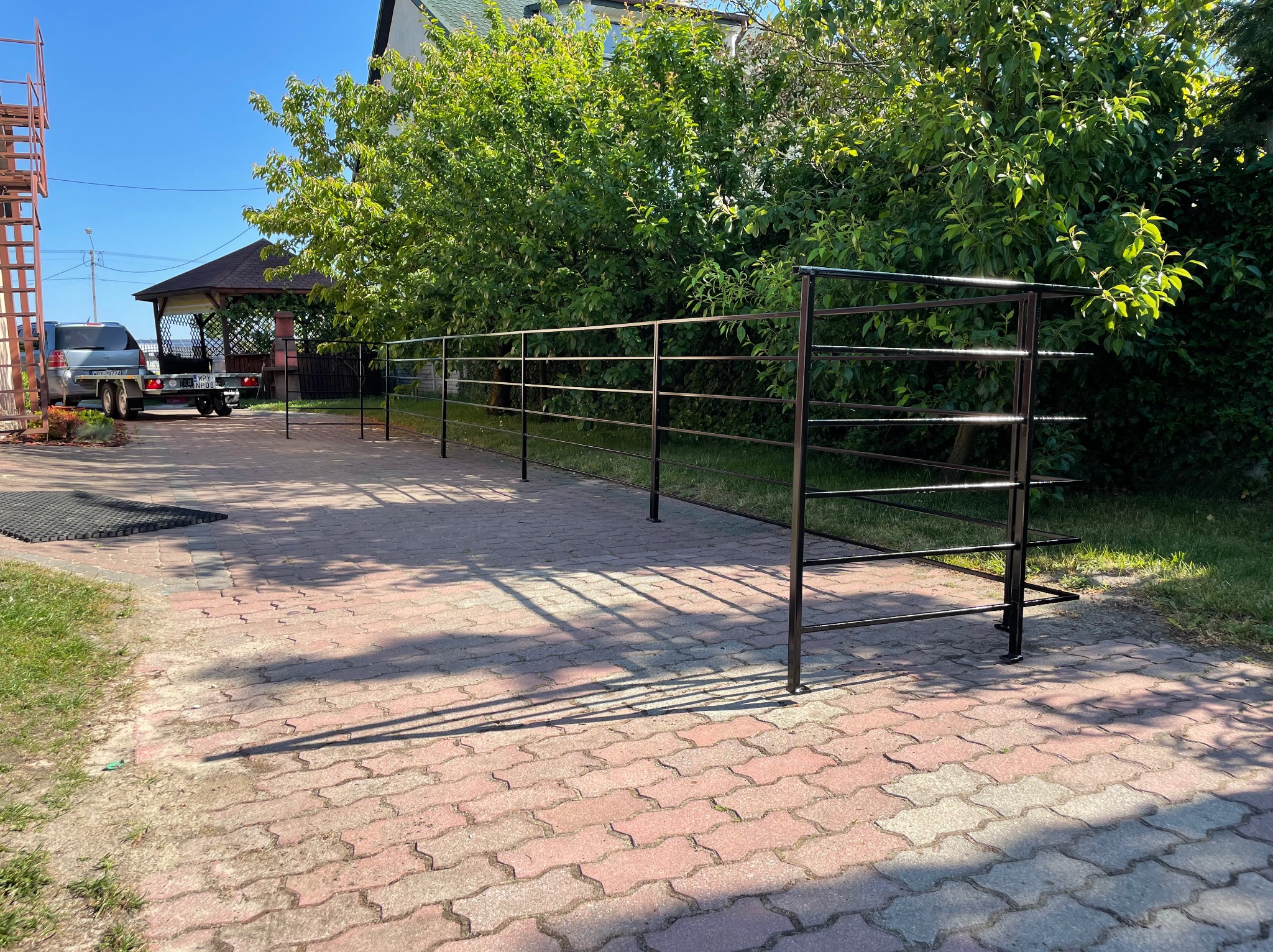 Czarne, metalowe ogrodzenie w trakcie montażu na posesji z kostki brukowej, widoczny fragment przyczepy samochodowej i altana ogrodowa w tle.