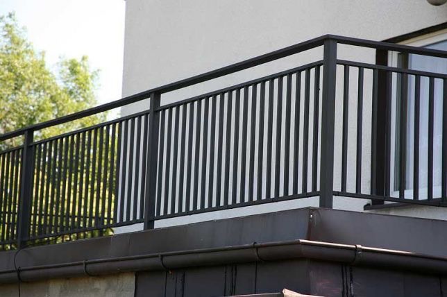 Czarna, metalowa balustrada balkonowa z pionowymi szczebelkami, widok z dołu, fragment elewacji budynku w tle.