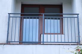 Balkon z prostą, metalową barierką w kolorze grafitowym, na tle białej elewacji i brązowych, przeszklonych drzwi balkonowych.