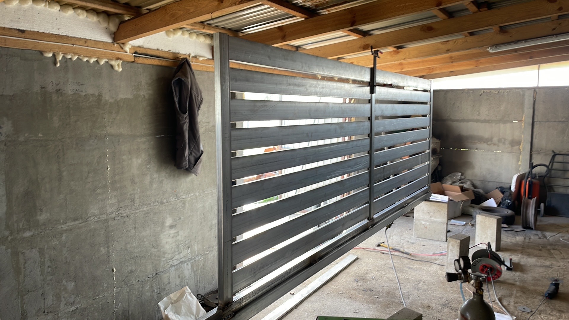 Surowy metalowy panel ogrodzeniowy z poziomymi szczeblami, zawieszony w warsztacie z betonowymi ścianami i narzędziami w tle, widoczne ślady spawania u dołu.