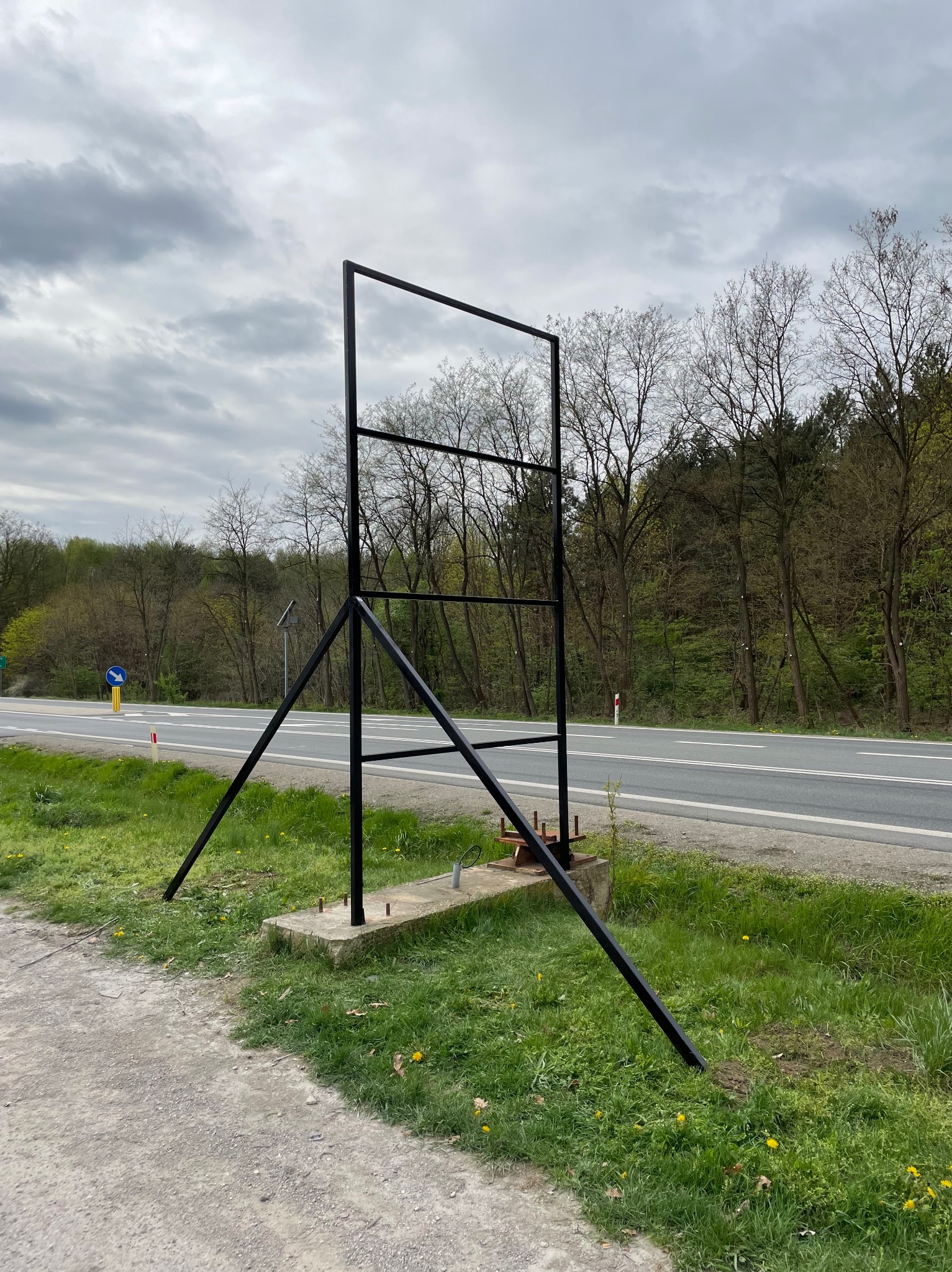 Surowa, czarna, metalowa konstrukcja stelaża reklamowego na betonowej podstawie, ustawiona przy drodze z widokiem na las i pochmurne niebo.