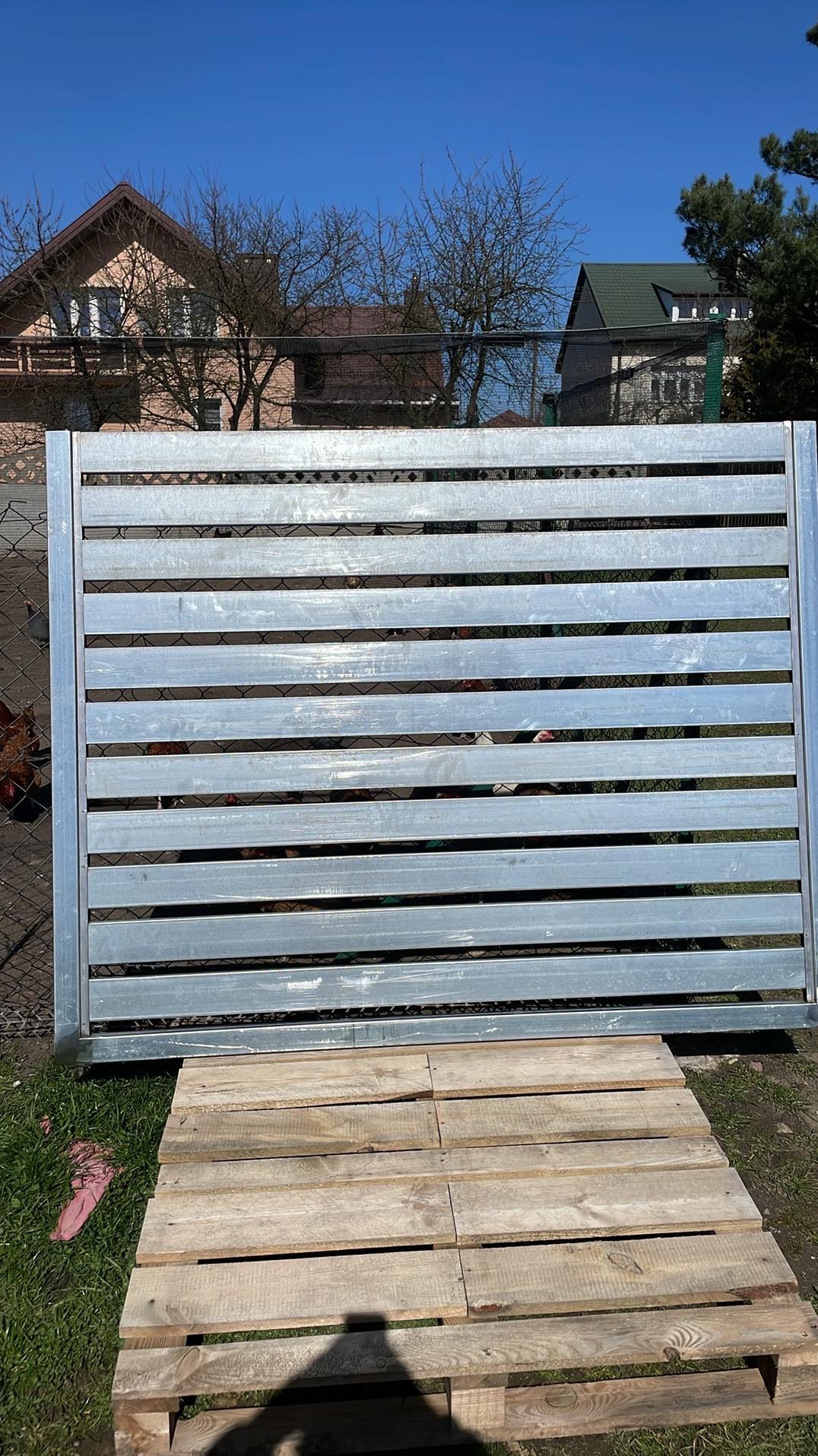 Element ogrodzenia z metalowych profili, ułożony na drewnianej palecie na trawie, w tle domy i błękitne niebo.