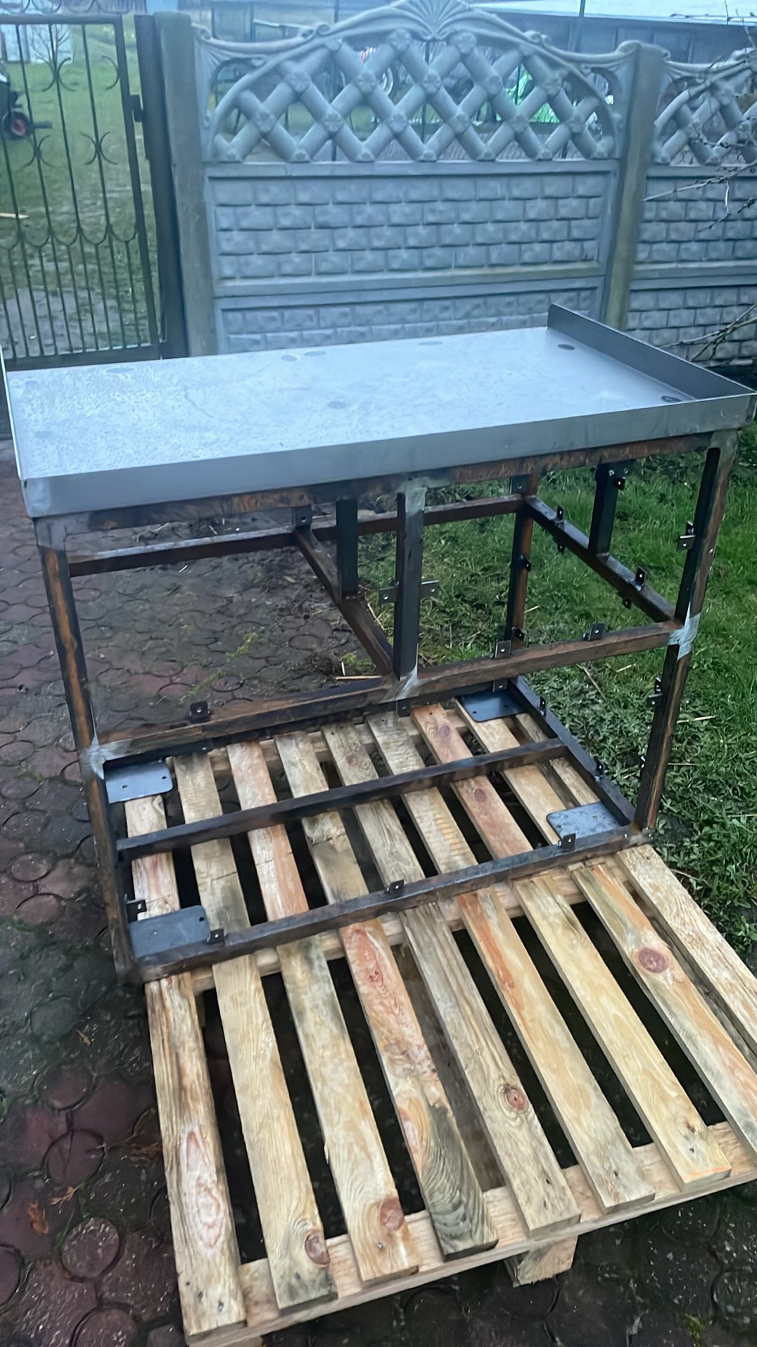 Stół roboczy ze stali nierdzewnej w trakcie budowy, umieszczony na drewnianej palecie, z widocznym ogrodzeniem betonowym w tle.