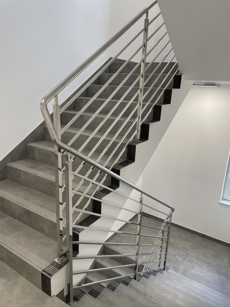 Schody wewnętrzne z szarymi płytkami i zabezpieczeniem w postaci metalowej konstrukcji z poziomych prętów, widok z góry na dół.