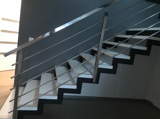 Schody z białymi stopniami i nowoczesną balustradą ze stali nierdzewnej z poziomymi prętami, zamontowaną na ciemnoszarej ścianie.