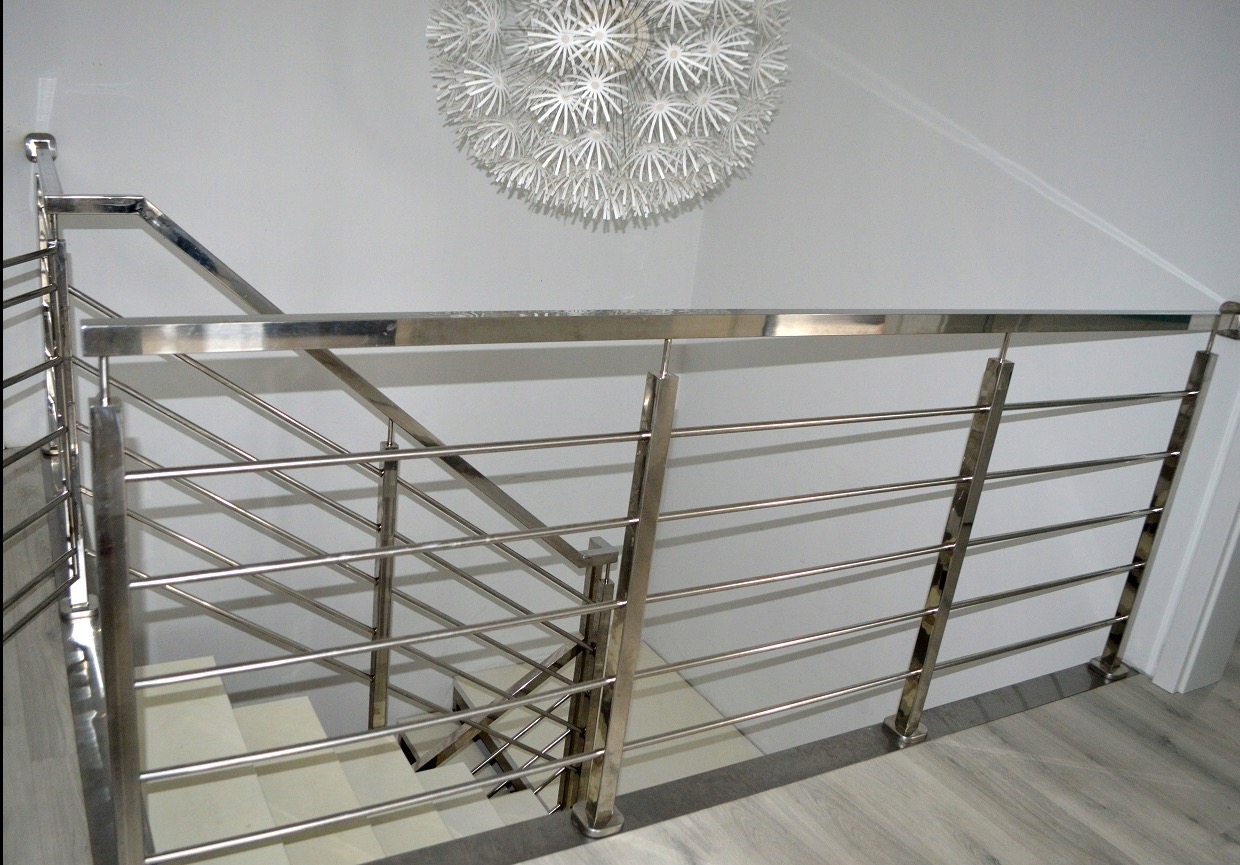 Błyszcząca, pozioma balustrada ze stali nierdzewnej z okrągłymi prętami, zamontowana na schodach wewnątrz domu, oświetlona przez nowoczesną lampę sufitową w kształcie kuli.