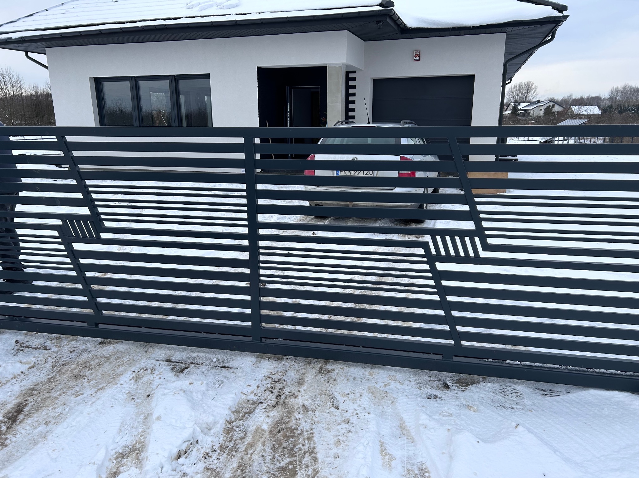 Nowoczesna, pozioma brama wjazdowa z szarego metalu o geometrycznym wzorze na tle zasypanego śniegiem podjazdu i białego domu z garażem.