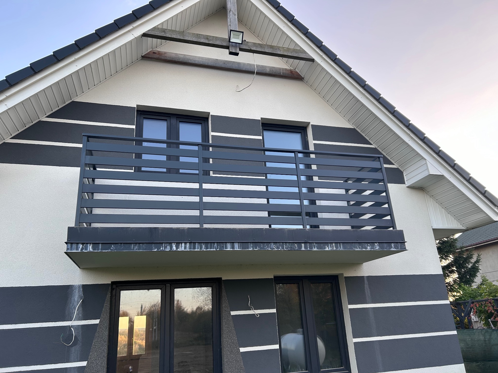 Balkon z grafitową balustradą poziomą, dopasowaną kolorystycznie do elewacji budynku z szarymi pasami, widok z dołu na fragment domu z oknami.
