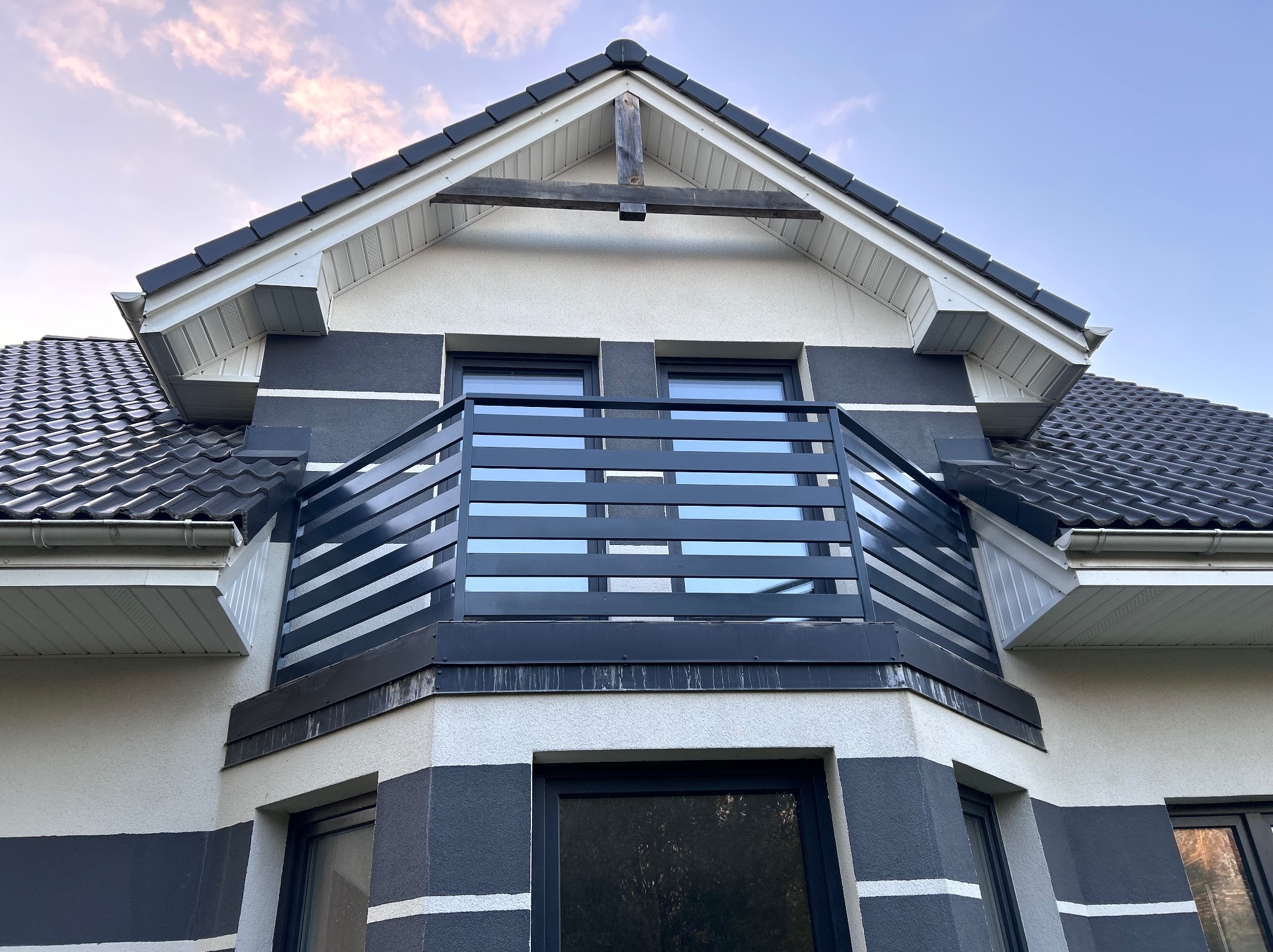 Nowoczesny balkon z poziomą, czarną balustradą na tle dwukolorowej elewacji domu z ciemną dachówką