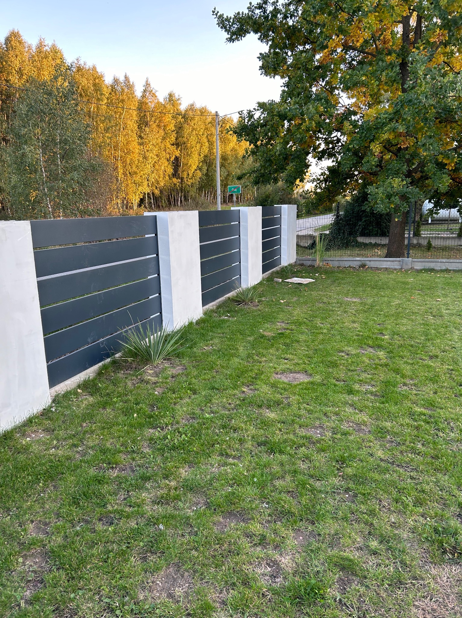 Nowoczesne ogrodzenie z poziomych, grafitowych paneli oddzielonych białymi, betonowymi słupkami na tle zielonego trawnika i jesiennych drzew.