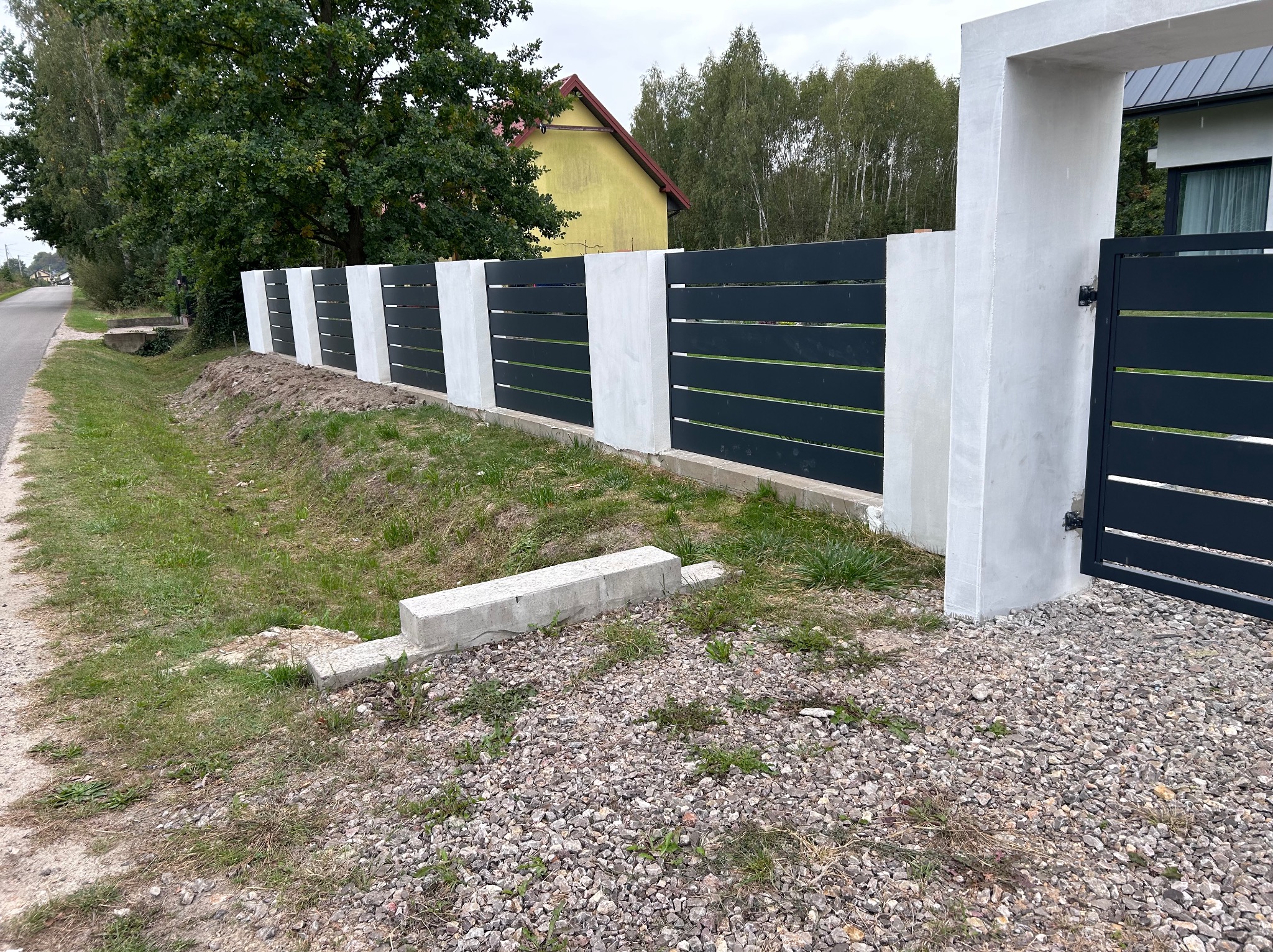 Nowoczesne ogrodzenie z poziomych, ciemnych paneli między białymi, betonowymi słupkami i bramą, widoczne na tle zieleni i domu.