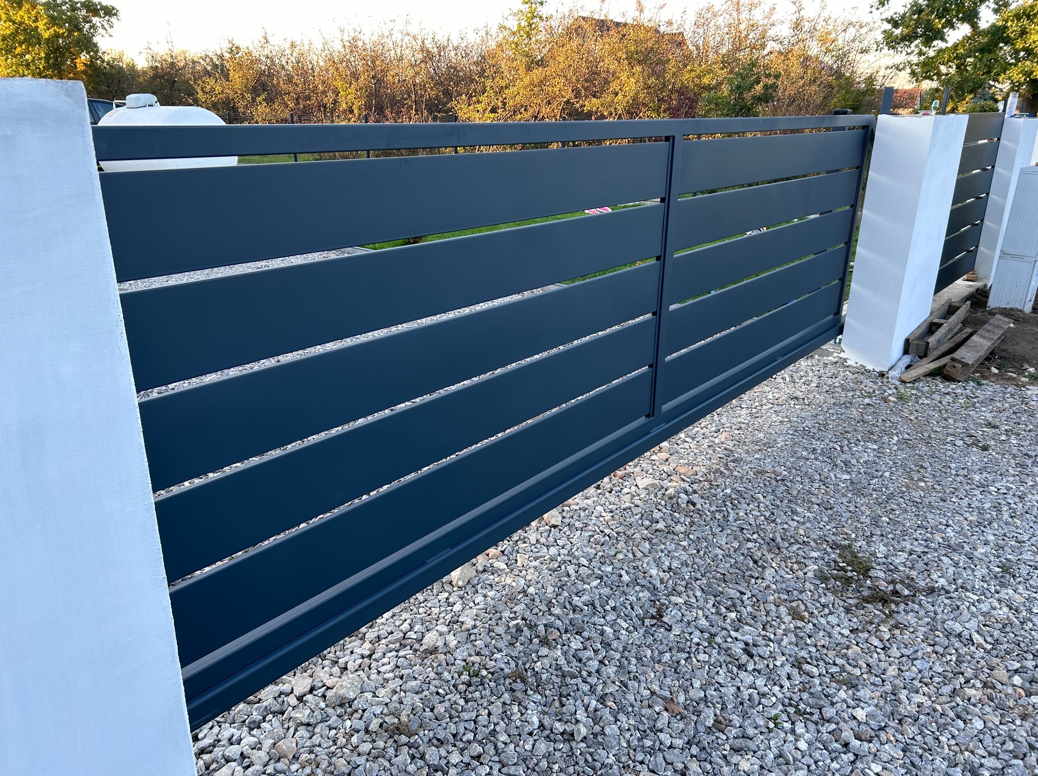 Nowoczesna, przesuwna brama w kolorze grafitowym z poziomymi panelami, osadzona między białymi, murowanymi słupkami na żwirowym podłożu.