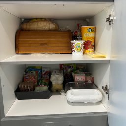 Małgorzata Kufta - Półka w szafce kuchennej wypełniona produktami spożywczymi, w tym pieczywem w chlebaku, płatkami śniadaniowymi, kremem orzechowym, herbatą i różnymi opakowaniami z przyprawami i dodatkami do potraw.