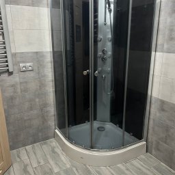 Małgorzata Kufta - Szara kabina prysznicowa z hydromasażem w narożniku łazienki wyłożonej szarymi płytkami imitującymi drewno.