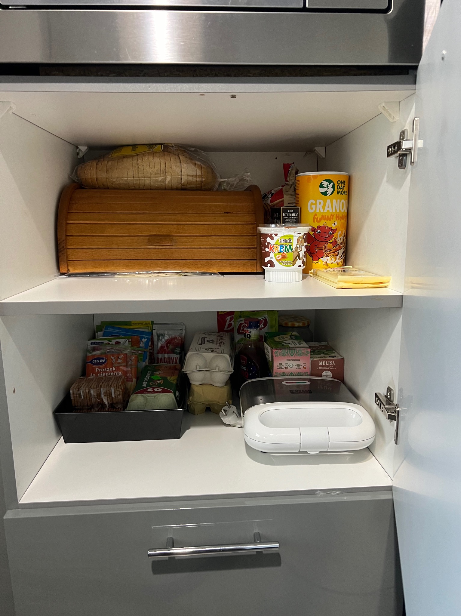 Półka w szafce kuchennej wypełniona produktami spożywczymi, w tym pieczywem w chlebaku, płatkami śniadaniowymi, kremem orzechowym, herbatą i różnymi opakowaniami z przyprawami i dodatkami do potraw.