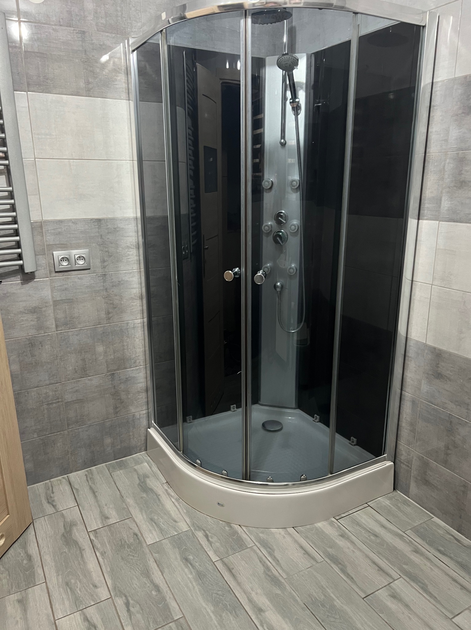 Szara kabina prysznicowa z hydromasażem w narożniku łazienki wyłożonej szarymi płytkami imitującymi drewno.