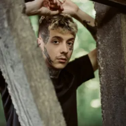 Portret młodego mężczyzny z tatuażami na twarzy, pozującego przy drewnianej konstrukcji na tle zieleni.