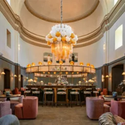 Eleganckie wnętrze baru z okrągłym barem, krzesłami barowymi z wzorzystymi siedziskami, r&oacute;żowymi fotelami i geometrycznym dywanem. Nad barem wisi dekoracyjny żyrandol z białych i złotych kul...