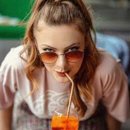 Percepcje Studio Paweł Aleksander Figiel - Młoda kobieta z rudymi włosami w okularach pije pomarańczowy napój przez papierową słomkę, patrząc prosto w obiektyw.