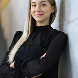 Percepcje Studio Paweł Aleksander Figiel - Portret uśmiechniętej blondynki w czarnej bluzce, opierającej się o ścianę z rękami skrzyżowanymi na piersiach.