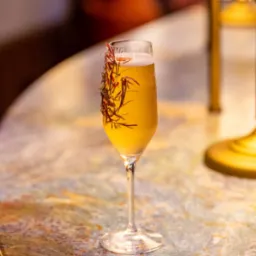 Kieliszek do szampana wypełniony złocistym napojem, ozdobiony suszonymi gałązkami, stoi na marmurowym blacie w ciepłym świetle lampki.