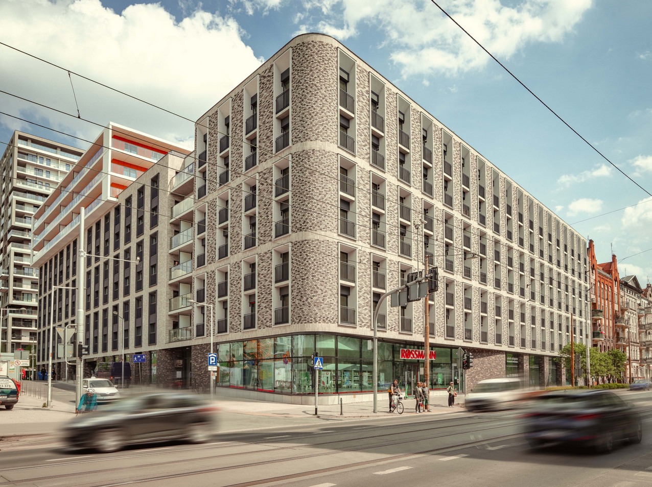 Nowoczesny budynek mieszkalny z ceglaną elewacją i balkonami, z widocznym parterem handlowym i przechodniami na chodniku.