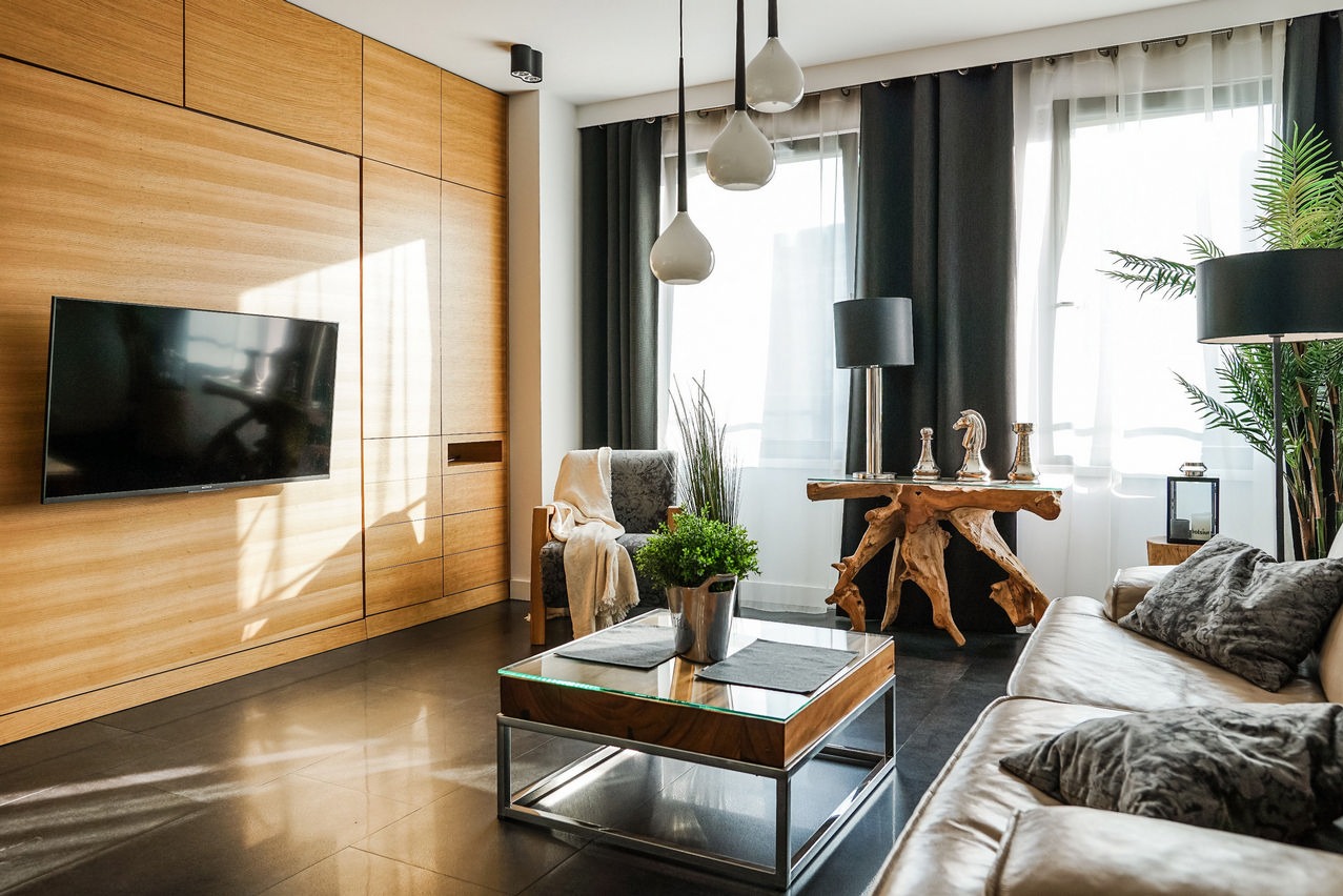 Nowoczesny salon w Krakowie z drewnianą zabudową ścienną, telewizorem, szklanym stolikiem kawowym i designerskimi lampami wiszącymi.