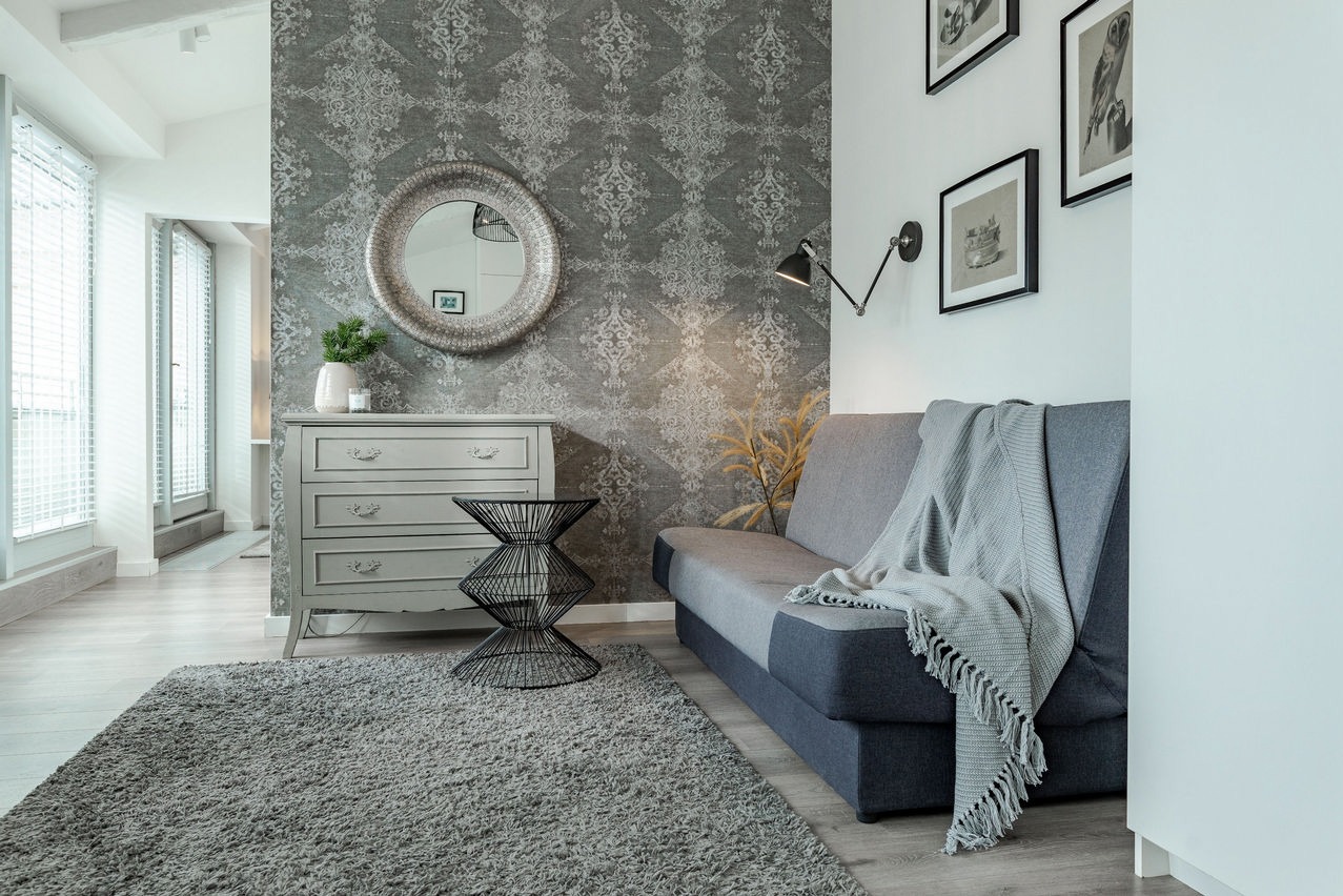 Stylowe wnętrze pokoju dziennego w odcieniach szarości z wzorzystą tapetą, komodą z okrągłym lustrem, szarą sofą z pledem i geometrycznym stolikiem kawowym. W tle widoczne okno balkonowe.