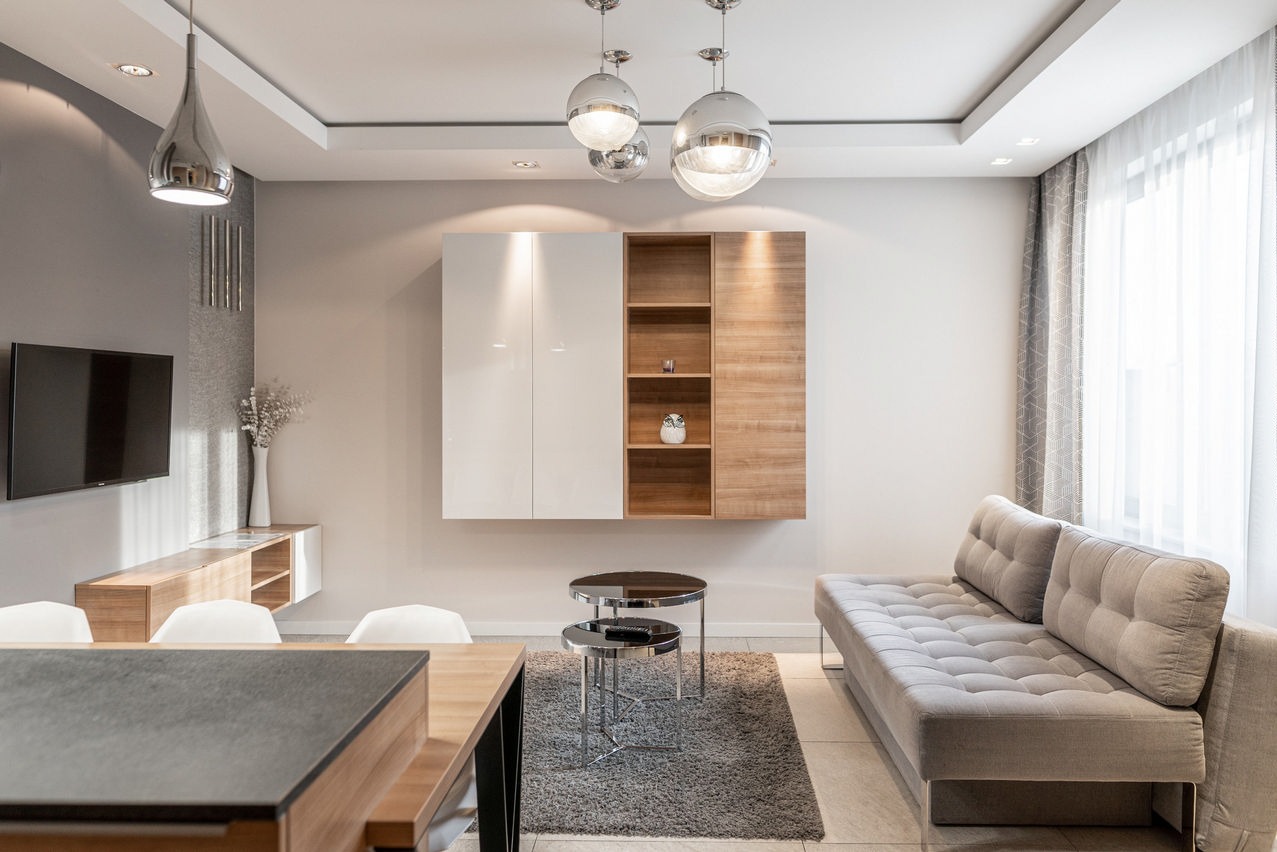 Jasne, nowocześnie urządzone wnętrze salonu z szarą sofą, drewnianymi meblami i okrągłymi lampami wiszącymi.