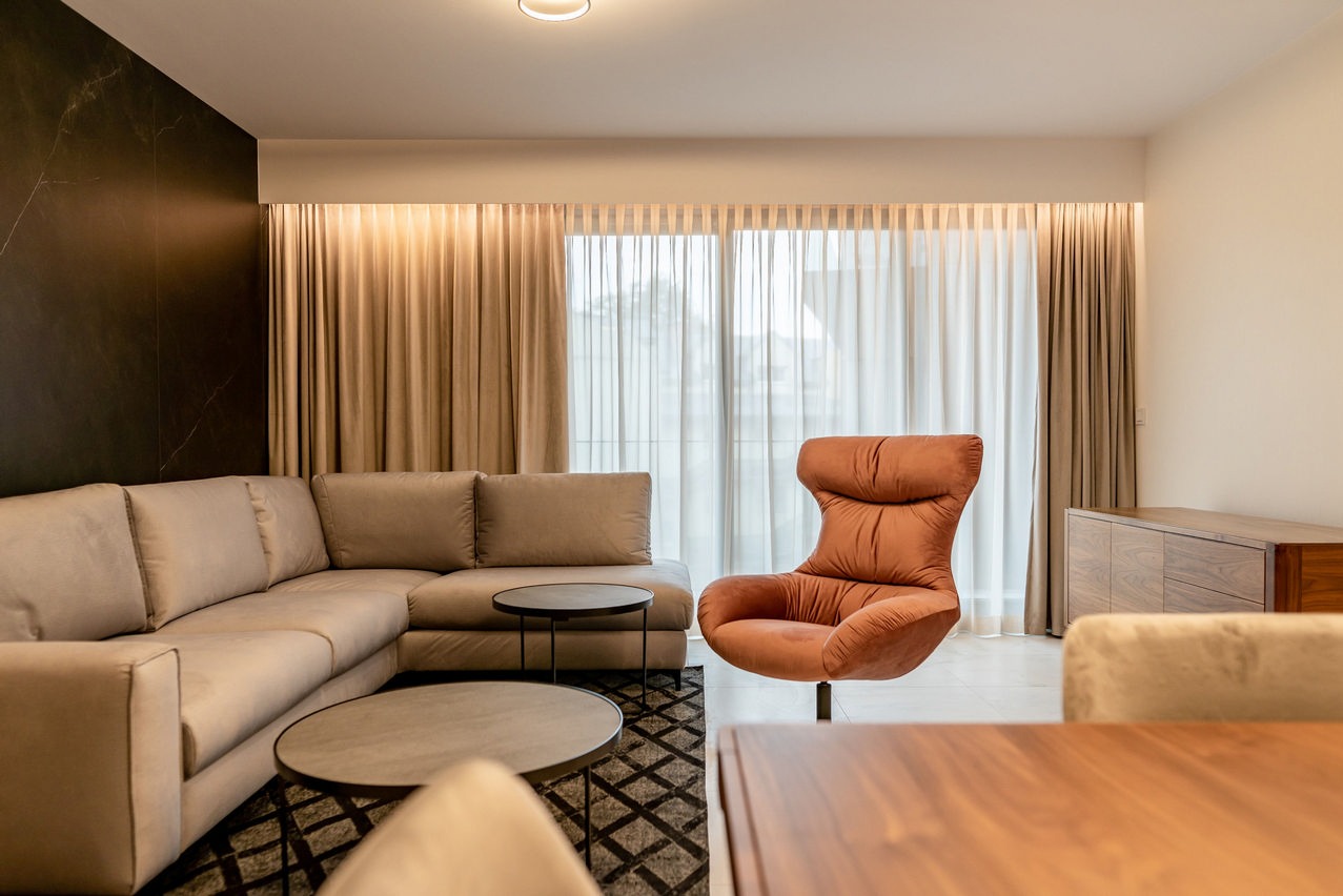 Jasny salon z narożną sofą, okrągłymi stolikami kawowymi, wzorzystym dywanem i fotelem w kolorze terakoty na tle okna z beżowymi zasłonami.