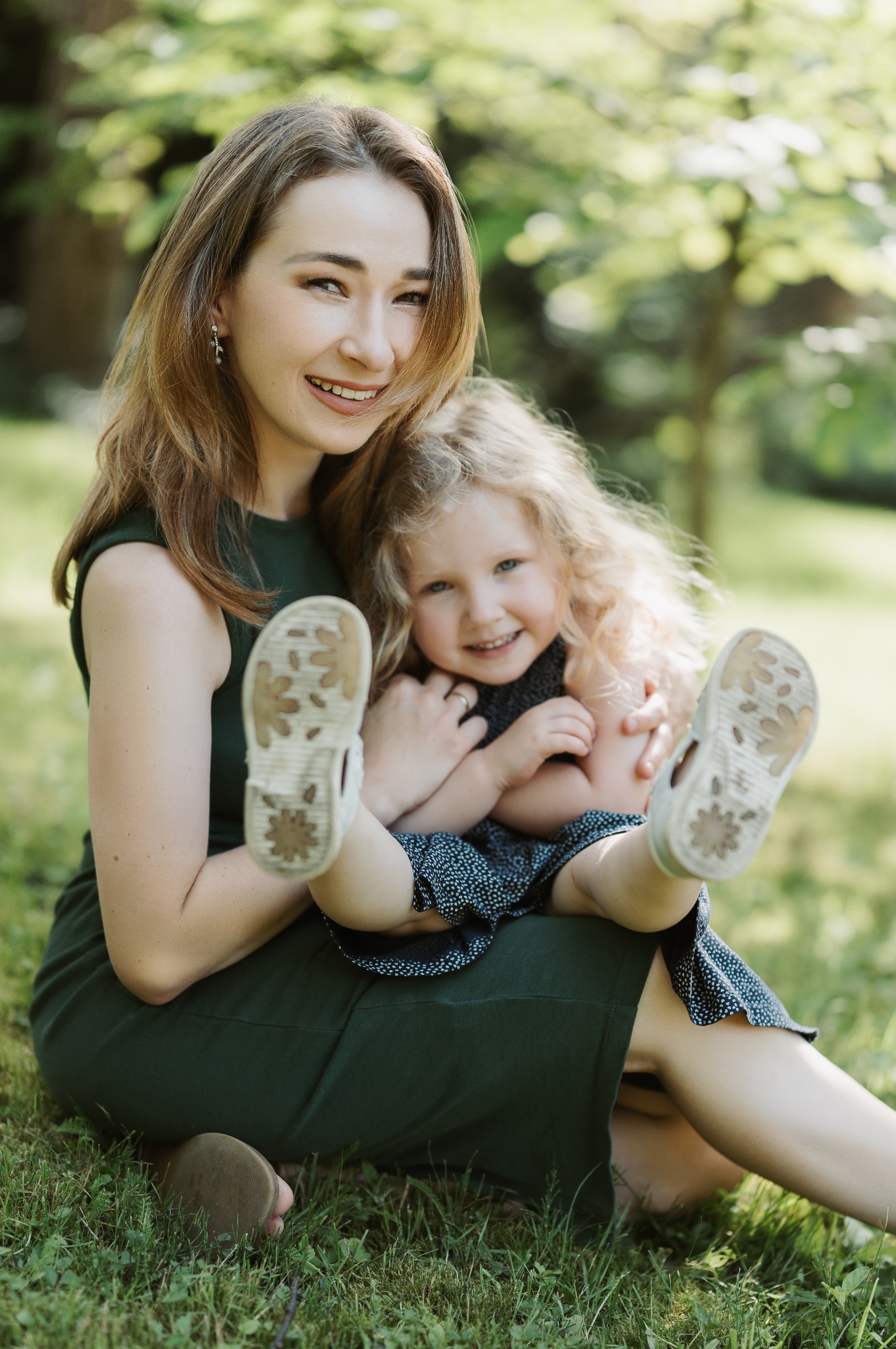 Uśmiechnięta mama w zielonej sukience trzyma na kolanach córeczkę z blond lokami, która wyciąga do przodu nóżki w sandałkach z widocznymi śladami użytkowania, siedząc na trawie w parku.
