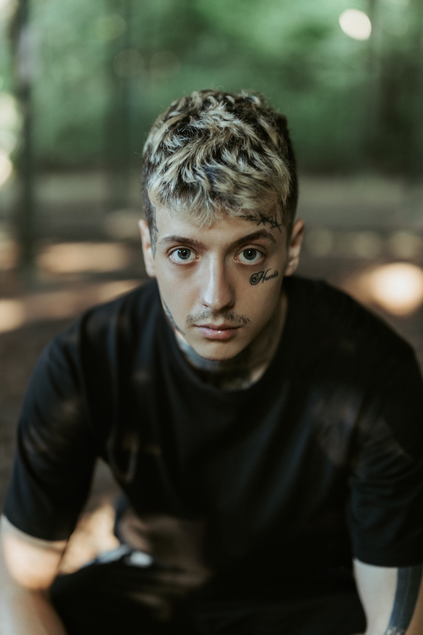 Młody mężczyzna z blond pasemkami, tatuażami na twarzy i szyi, ubrany w czarny T-shirt, siedzący na tle zieleni.