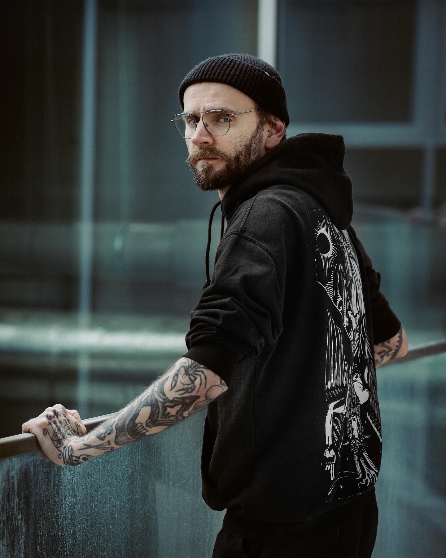 Mężczyzna w czarnej bluzie z kapturem i czapce, z tatuażami na rękach, oparty o metalową barierkę, spogląda w bok na tle szklanej fasady budynku.