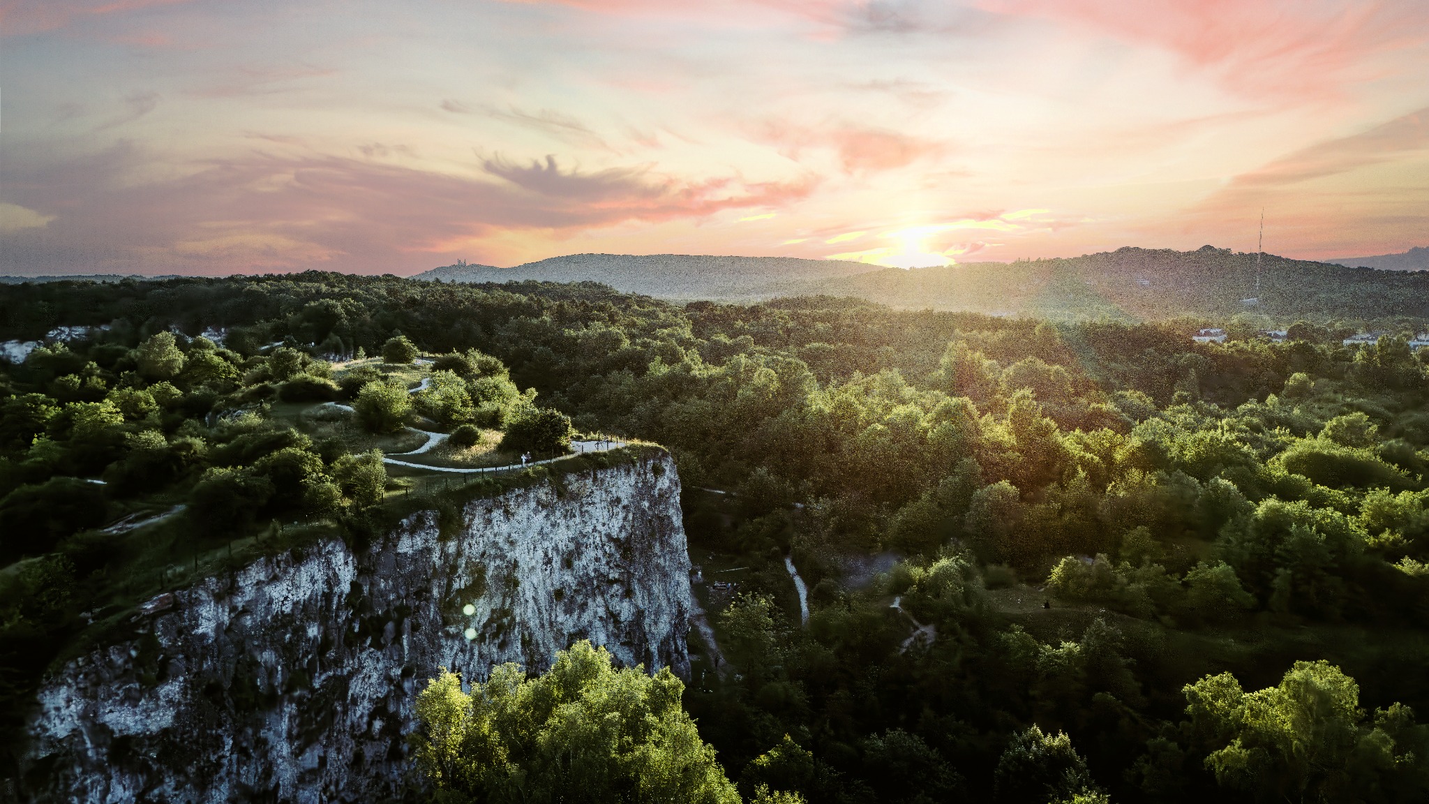 Panoramiczny widok z lotu ptaka na wapienny klif porośnięty drzewami, z ścieżką na szczycie i sylwetkami spacerujących osób, w tle zalesione wzgórza i zachodzące słońce z różowawymi chmurami.
