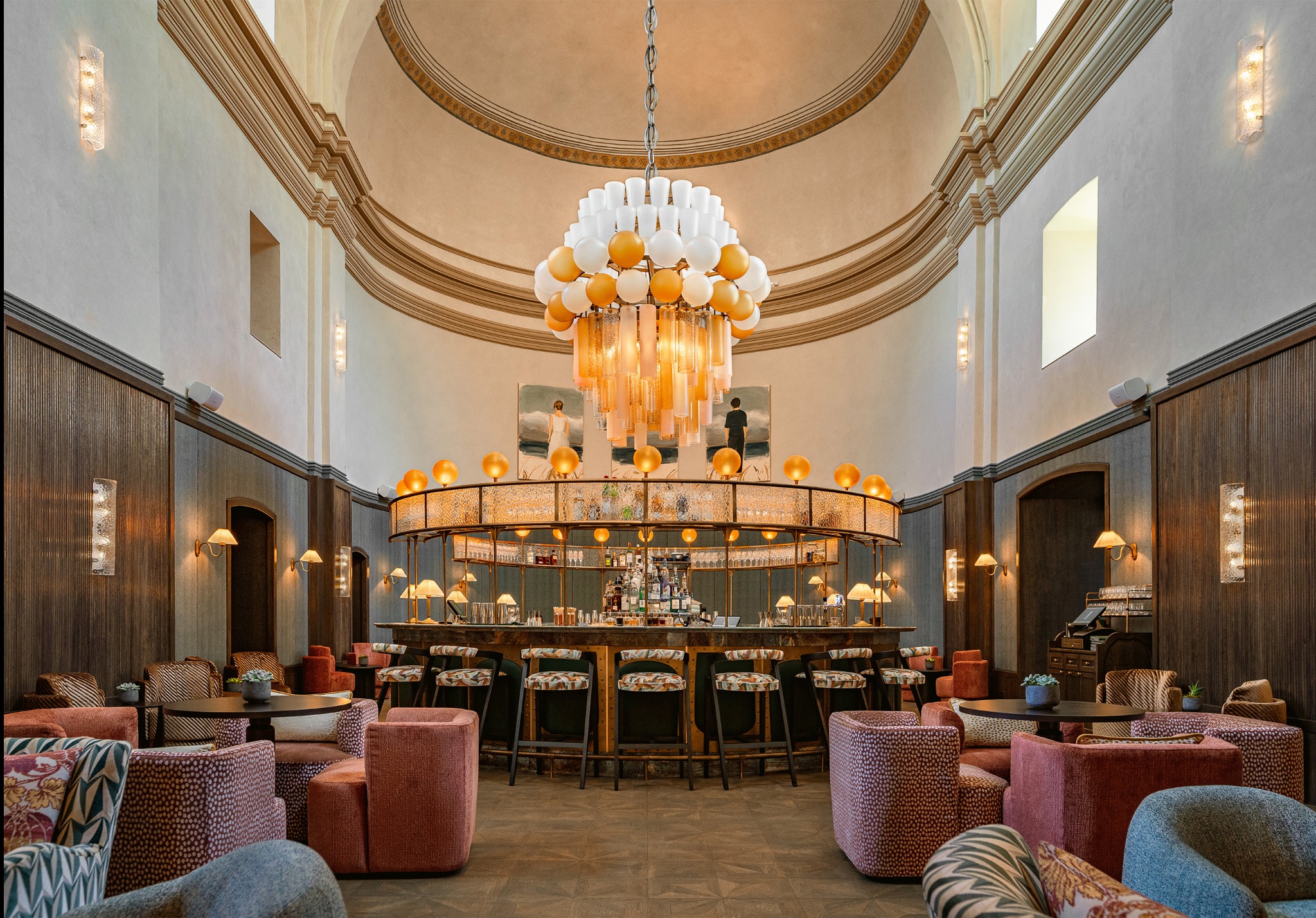 Eleganckie wnętrze baru z okrągłym barem, krzesłami barowymi z wzorzystymi siedziskami, różowymi fotelami i geometrycznym dywanem. Nad barem wisi dekoracyjny żyrandol z białych i złotych kul...