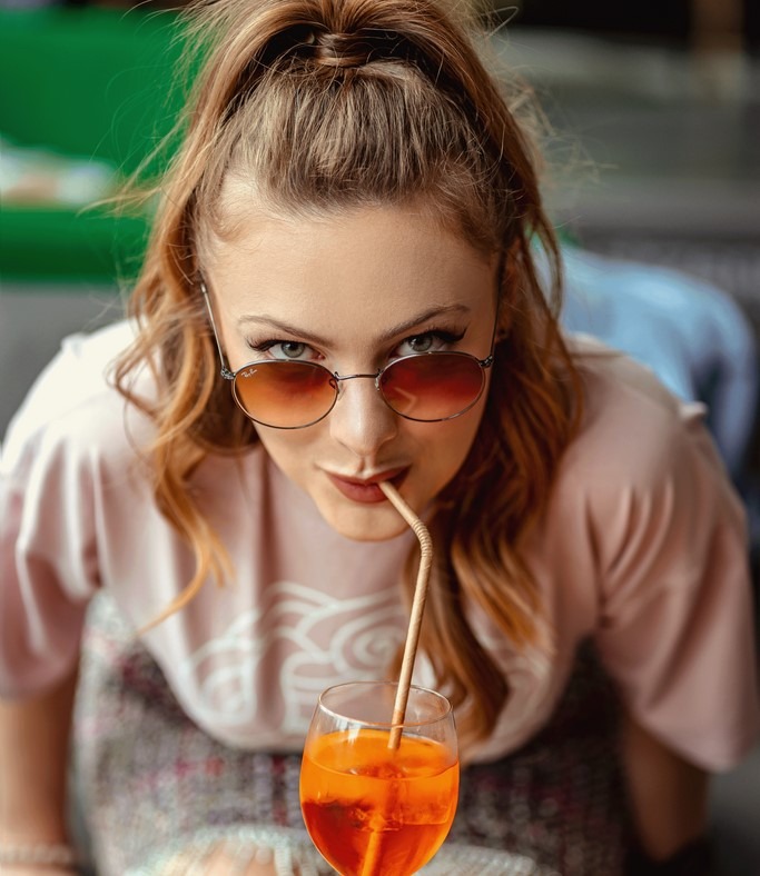 Młoda kobieta z rudymi włosami w okularach pije pomarańczowy napój przez papierową słomkę, patrząc prosto w obiektyw.