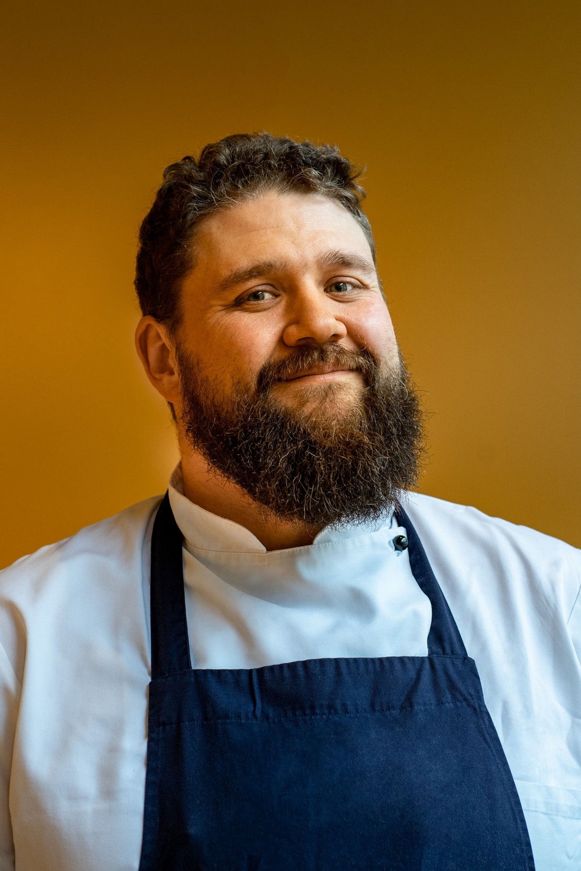 Portret uśmiechniętego kucharza z brodą, w białym uniformie i granatowym fartuchu na tle jednolitego, ciepłego koloru.