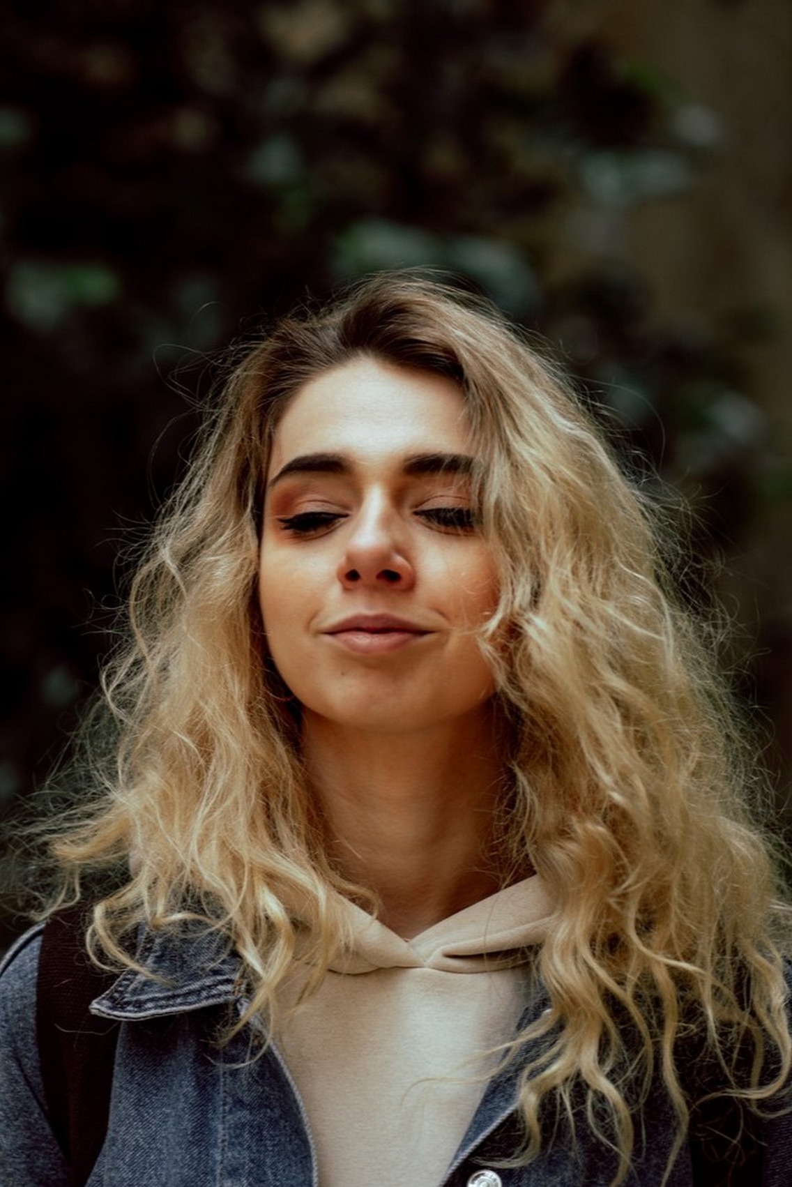 Portret młodej kobiety z blond, kręconymi włosami, ubrana w dżinsową kurtkę i beżową bluzę z kapturem, z delikatnym makijażem oczu i zamyślonym wyrazem twarzy na tle ciemnego, zamazanego tła.