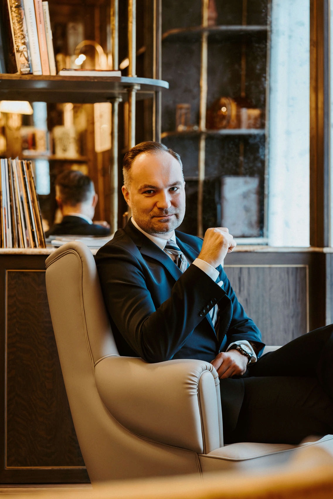 Mężczyzna w garniturze siedzi w fotelu, z półkami i książkami w tle, z odbiciem w lustrze.