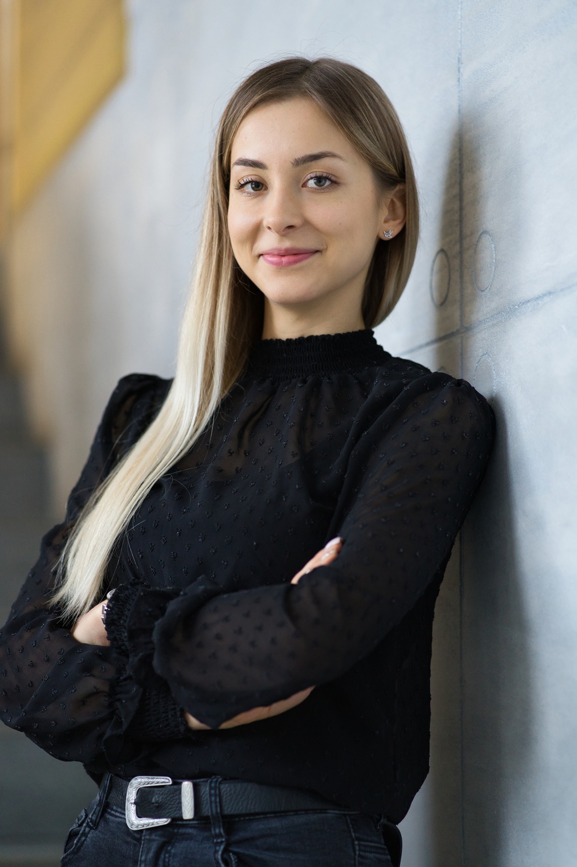 Portret uśmiechniętej blondynki w czarnej bluzce, opierającej się o ścianę z rękami skrzyżowanymi na piersiach.
