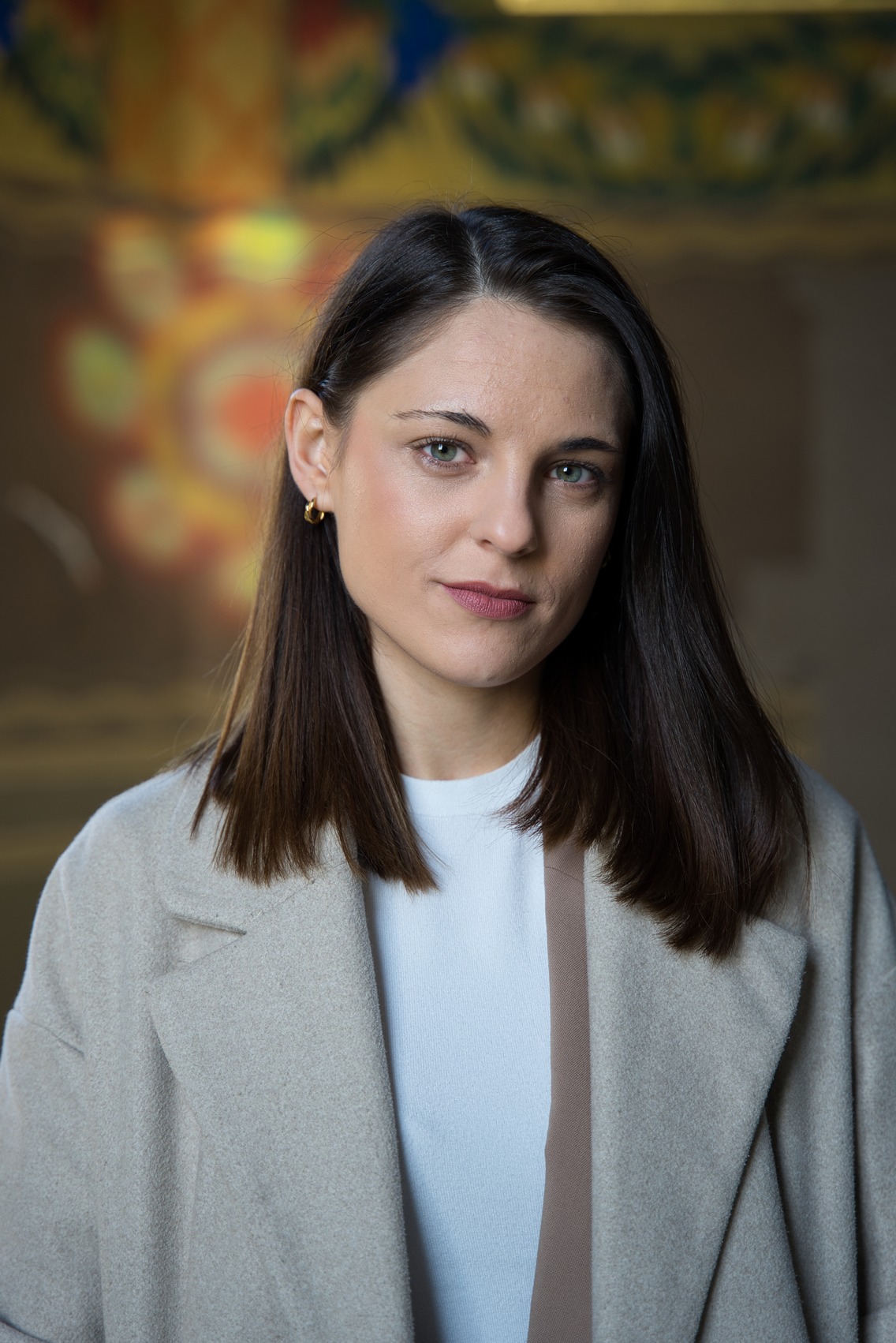 Portret kobiety w jasnym płaszczu na rozmytym tle w Krakowie. Uwagę zwracają zielone oczy i złote kolczyki.