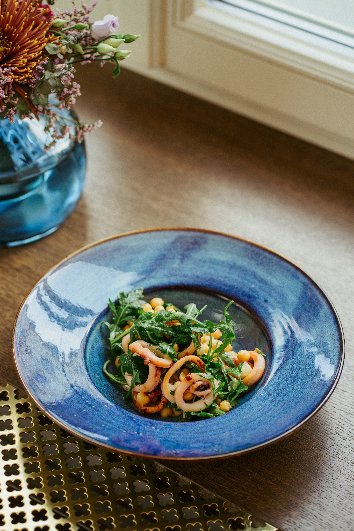 Stylizowana sałatka z kalmarami, ciecierzycą i rukolą na niebieskim, ceramicznym talerzu z rustykalnym wykończeniem, obok wazon z kwiatami i złota podkładka z wzorem koniczyny na drewnianym stole.